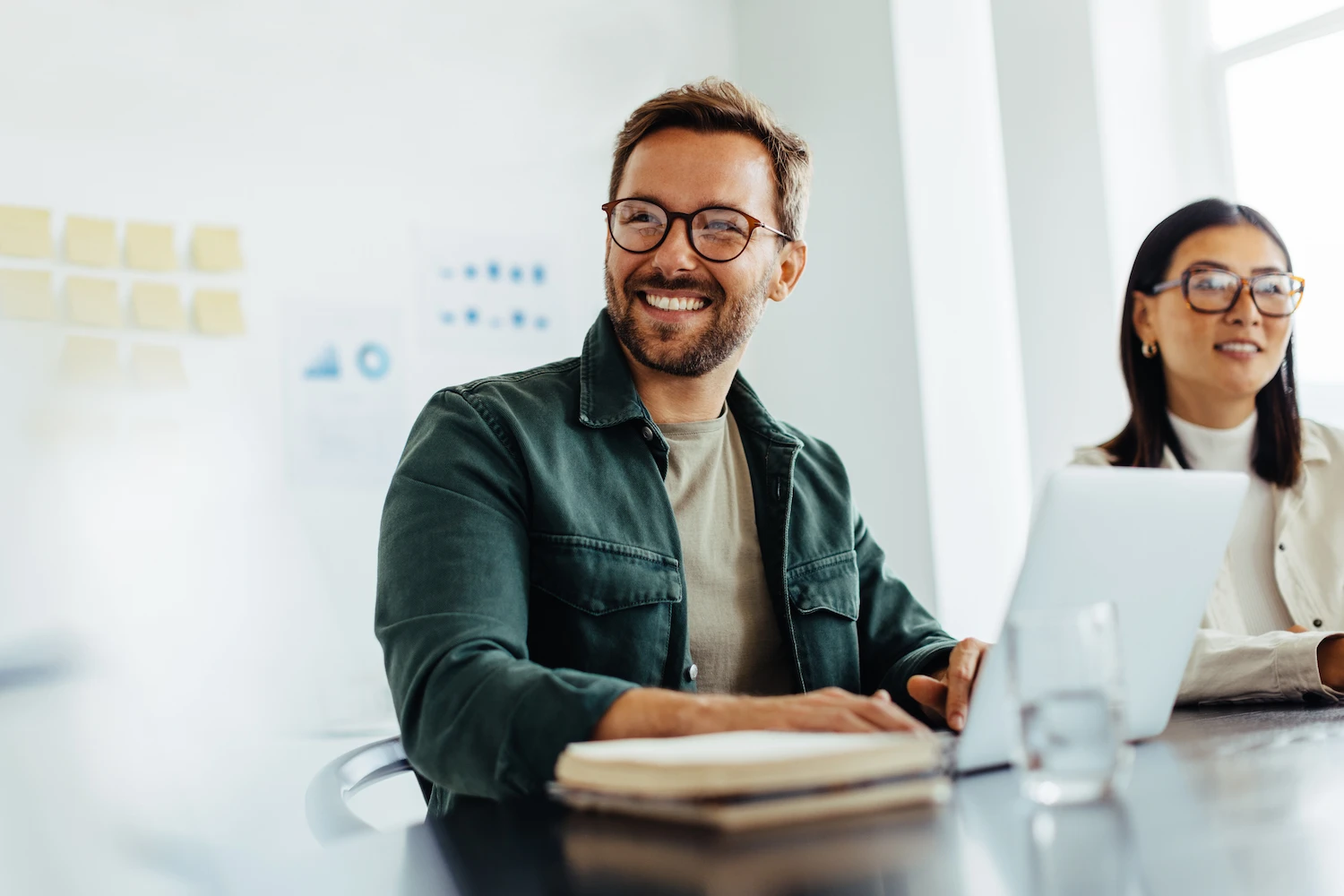 CoworkersChat-Bright Smiling man with glasses in a green shirt working on a laptop at a bright, modern office desk with a colleague and blurred wall notes in the back.