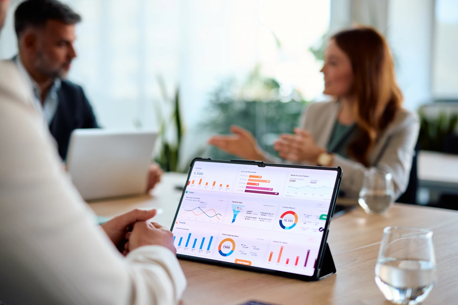 Tablet-Stats Tablet displaying a business analytics dashboard with charts and financial data in a bright, professional boardroom meeting with blurred colleagues.
